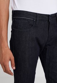 Jeans en denim foncé avec une coupe près du corps, dotés d'une configuration classique à cinq poches et de coutures contrastantes le long des seams.