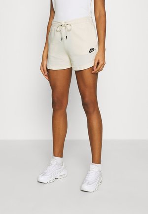 Person iført beige Nike-shorts med snøre, hvide sokker, hvide Nike-sneakers og en hvid ærmeløs top, som står på en grå gulv.