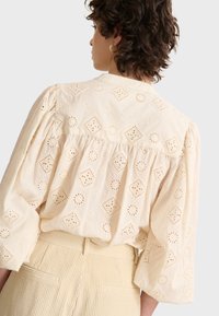 Crème blouse met bloemenoog-in-gaaspatroon, pofmouwen en geplooide achterkant. Gecombineerd met lichtgekleurde geribde broeken.