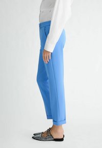 Světle modré kalhoty na míru s zužujícím střihem a ohrnutými manžetami, spárované s černými slip-on botami s bílými hroty.