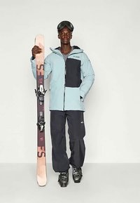 Svetlo modra smučarska jakna s črnim panelom, črne snežne hlače in črne smučarske čizme; drži rožnate in črne smuči; zaščitna očala na glavi.