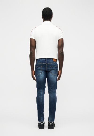 Homme aux cheveux courts portant une chemise blanche à manches courtes, un jean bleu déchiré et des baskets noires avec des logos de feuilles blanches, debout de dos.