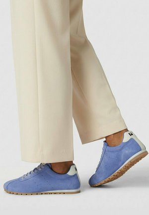 Persoon die lichtbeige broek en blauwe suède sneakers met witte hielaccenten en gumzolen draagt, staande op een lichtgrijze vloer.
