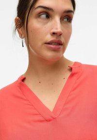 Blusa coral de cuello en V con una textura suave y un sutil detalle de corte en el escote. La vista de cerca resalta la costura y la suavidad de la tela.