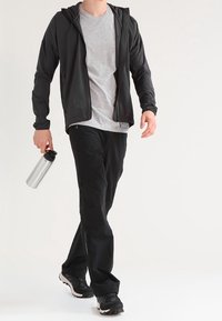 Svart lättviktsjacka med dragkedja, grå t-shirt med rund hals, svarta sportbyxor och sneakers. Person som håller i en silverfärgad vattenflaska.