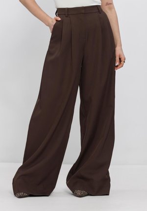 Woman wearing high-waist, wide-leg dark brown trousers over leopard print pointed shoes, with one hand in pocket, standing against plain background.