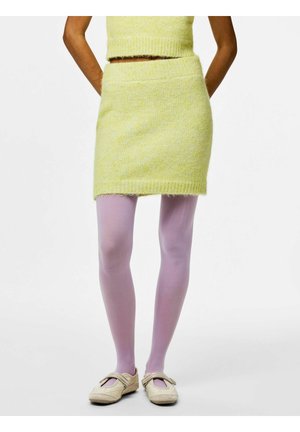 Person iført et gult, pelsagtigt skørt og top, lavendelfarvede strømpebukser og cremefarvede Mary Jane-sko, stående med hænderne bag ryggen.