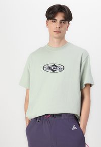 Light green cotton t-shirt with short sleeves and a round neckline. Features a black and blue "Rip Curl" logo on the front.