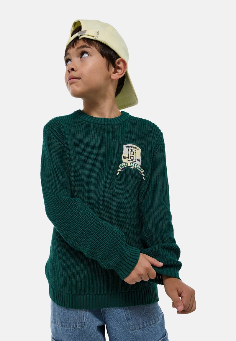Pull en tricot vert avec un col rond, des poignets côtelés et un écusson avec une lettre et du texte. Porté avec un short en denim bleu clair.