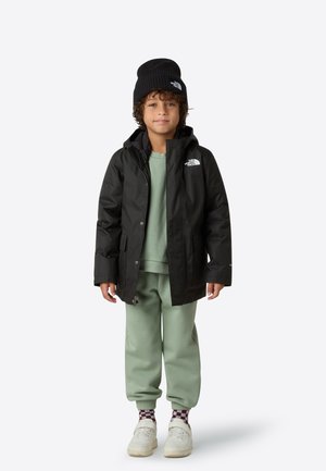 Giacca nera impermeabile con cappuccio, felpa verde sotto, pantaloni della tuta verde chiaro, berretto nero e scarpe bianche con calzini a fantasia.