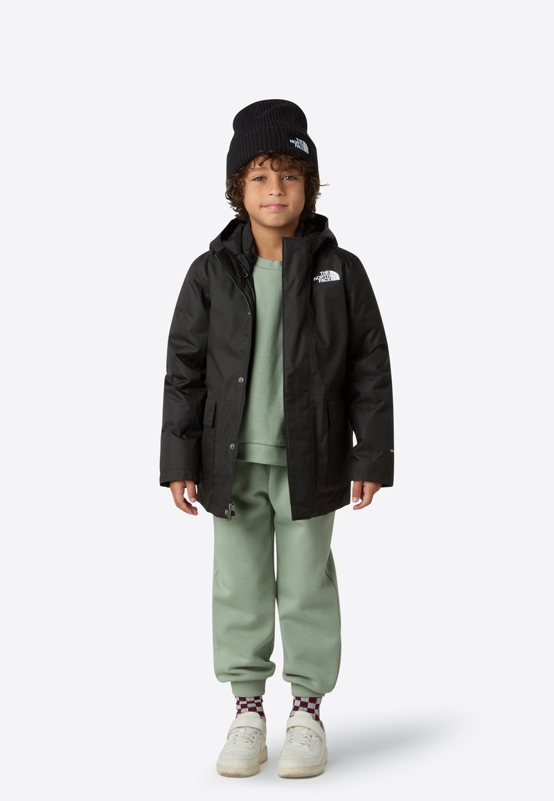 Černá nepromokavá bunda s kapucí, zelená mikina pod ní, světle zelené tepláky, černá čepice a bílé tenisky s vzorovanými ponožkami.