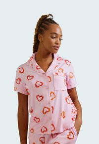Pink pajama set with short sleeves, featuring red and orange heart patterns, a breast pocket, and a relaxed fit. Lightweight fabric texture.