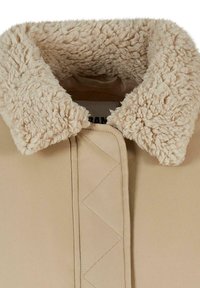 Giacca beige con un colletto texturizzato e soffice. Il colletto è di colore crema con una superficie morbida e pelosa. La parte anteriore presenta un motivo trapuntato visibile.