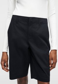 Άτομο που φοράει μαύρα σορτς μέχρι το γόνατο και λευκό μακρυμάνικο τοπ, στέκεται με τα χέρια χαλαρά στα πλάγια μπροστά σε μια απλή φόντο.