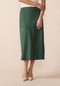 Jupe midi en satin vert avec une texture lisse, une forme légèrement évasée et un design uni. Portée avec des escarpins beiges à bout pointu.