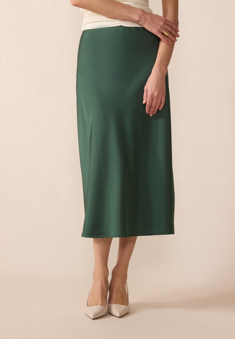 Jupe midi en satin vert avec une texture lisse, une forme légèrement évasée et un design uni. Portée avec des escarpins beiges à bout pointu.