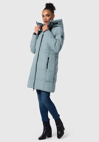 Lichtblauwe pufferjas met capuchon, voorzien van een rits aan de voorkant, zwarte accenten en een gewatteerd ontwerp. Model draagt donkere jeans en zwarte hakken.
