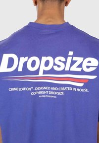 Lila Baumwoll-T-Shirt mit großen weißen "Dropsize"-Text auf der Rückseite, verziert mit roten und blauen Akzenten. Der Text enthält Markendetails und Copyright.