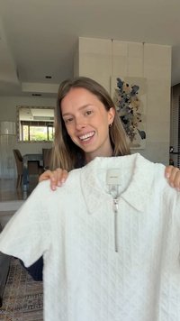 Young woman smiling indoors holding up a white textured short-sleeve shirt with a front zipper and collar.
