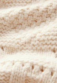 Světle béžová pletenina s měkkým, texturovaným povrchem a viditelnými švy, která vykazuje útulný, chunky design.
