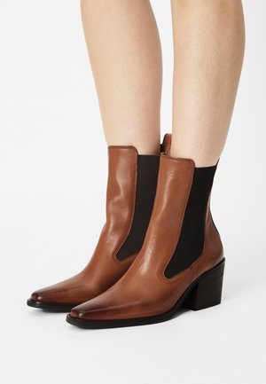 Bottines en cuir marron avec des panneaux élastiques sur les côtés, bout pointu et talon empilé. Texture lisse et design épuré.