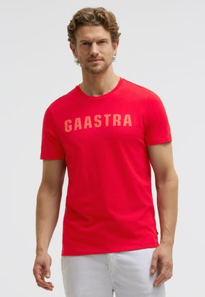 Mann mit hellbraunen lockigen Haaren, der ein leuchtend rotes T-Shirt mit der Aufschrift "GAASTRA" auf der Brust und weiße Hosen trägt, steht vor einem schlichten Hintergrund.