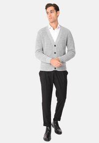 Cardigan grigio a coste con scollo a V, caratterizzato da tre bottoni neri, abbinato a pantaloni neri e stivali neri con lacci. Camicia bianca sotto.