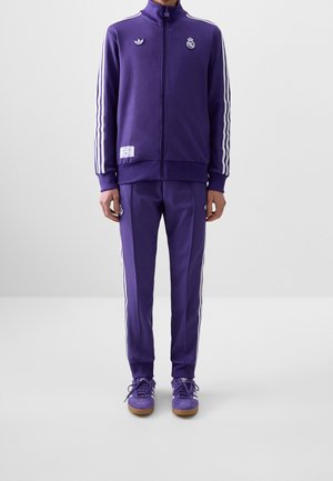 Homme portant un survêtement violet Adidas avec des bandes blanches, des logos du Real Madrid et des baskets Adidas violettes assorties.