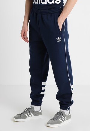 Mørkeblå Adidas træningsbukser med hvide striber på siden og trefoil-logo, båret med grå Adidas sneakers og hvide sokker.