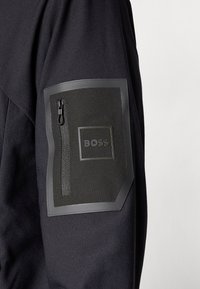 Μαύρο μανίκι μπουφάν με υφασμάτινη υφή, ορθογώνιο μαύρο λάκκο και τσέπη με φερμουάρ που φέρει πράσινο λογότυπο "BOSS".