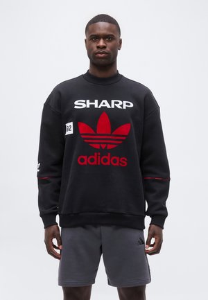 Muškarac nosi crnu Adidas majicu s crvenim logotipom i tekstom "SHARP", u kombinaciji sa sivim Adidas šortsevima, stoji ispred jednobojne pozadine.