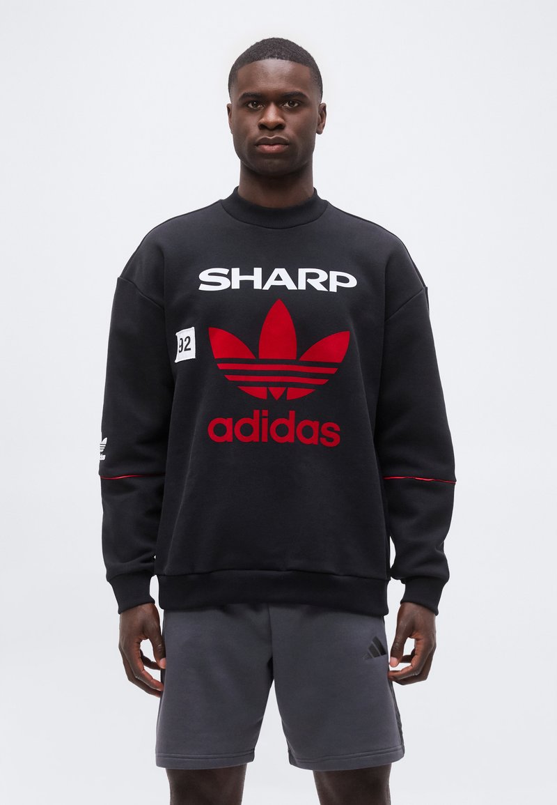 Άνδρας με μαύρο φούτερ Adidas με κόκκινο λογότυπο και κείμενο "SHARP", συνδυασμένο με γκρι σορτς Adidas, στέκεται μπροστά σε απλό φόντο.