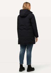 Abrigo acolchado azul marino con capucha, de corte recto, de longitud media y textura suave. Combinado con jeans ajustados azules y botas negras.
