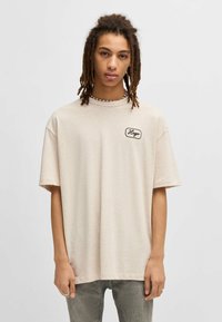 Beige oversized t-shirt van zachte katoen, met een zwart geborduurd logo op de borst. Korte mouwen en ronde halslijn.