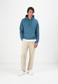 Felpa teal con tasca a marsupio e un piccolo logo giallo sul petto, abbinata a pantaloni beige chiaro e sneaker bianche.