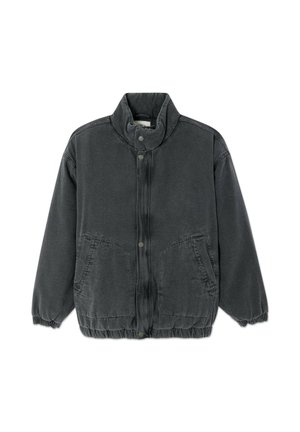 Jeune homme aux cheveux foncés portant une veste noire zippée, un pantalon bleu et des chaussures noires, debout les mains dans les poches devant un fond uni.