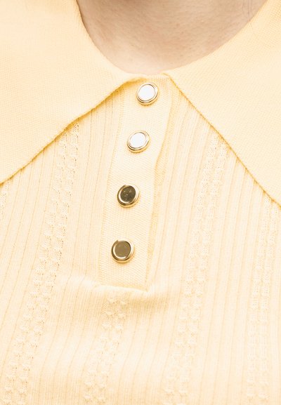 Κοντινό πλάνο ενός κίτρινου πλεκτού τοπ με φαρδύ γιακά και τέσσερα κουμπιά με χρυσές λεπτομέρειες σε κάθετη ριμπ ύφανση.