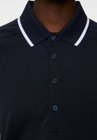 Marinefarbendes Poloshirt mit kontrastierendem weißen Kragen. Verfügt über eine Knopfleiste und strukturierte Stoffoberfläche. Mehrere Knöpfe sind auf der Vorderseite sichtbar.