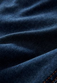 Sötétkék farmeranyag látható textúrával és finom redőkkel, amely finom szövést és narancssárga varrást sejtet jobb alsó sarokban.