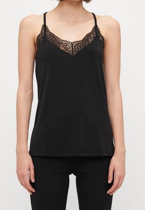 Kvinde iført sort camisole med blonder og tynde stropper samt sorte bukser, stående mod en ensfarvet lys baggrund.