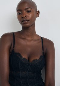 Fekete csipke bodysuit, amely finom virágmintázattal, állítható pántokkal és testéhez simuló, texturált, áttetsző dizájnnal rendelkezik.