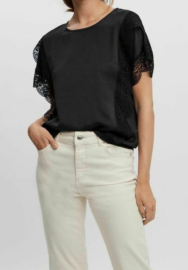 Femme portant un haut noir à manches courtes avec des détails en dentelle et un pantalon beige clair taille haute, une main dans la poche, sur un fond uni.