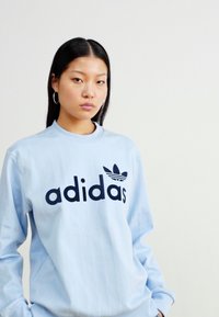 Sudadera Adidas de color azul claro con textura acanalada, que presenta un gran logo negro y un diseño de trébol en la parte delantera. Mangas largas y cuello redondo.