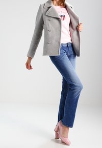 Cappotto grigio doppiopetto sopra una camicia rosa con logo, abbinato a jeans blu e scarpe con tacco quadrato rosa. Uno sfondo semplice accentua il focus.