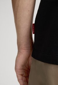 T-shirt noir avec une étiquette rouge visible portant la mention "Joop! Jeans". La main à côté est de teint moyen, mettant en valeur la texture de la peau.