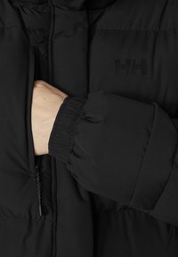Černá péřová bunda s kapsou na zip a elastickými manžetami. Materiál je hladký s matným povrchem, na kterém se nachází jemné logo.
