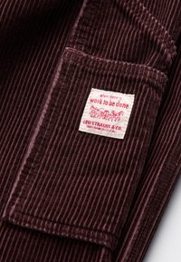 Tejido de pana marrón con costillas verticales, con una etiqueta blanca que muestra "cuando hay trabajo que hacer" y el logo de Levi Strauss.