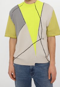 Homme portant un gilet en tricot à motifs en patchwork avec des sections jaunes néon, beige et grises sur une chemise jaune-vert à manches courtes et un jean bleu.