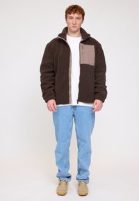 Brown fleece jacket with a zippered pocket in light brown, collar up, worn over a white shirt, paired with blue jeans and beige shoes.