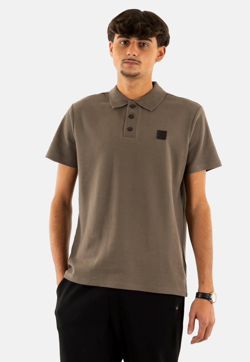 Camisa polo de manga corta en verde oliva, con tejido texturizado, cierre de tres botones y un pequeño parche de logo negro en el lado izquierdo del pecho.